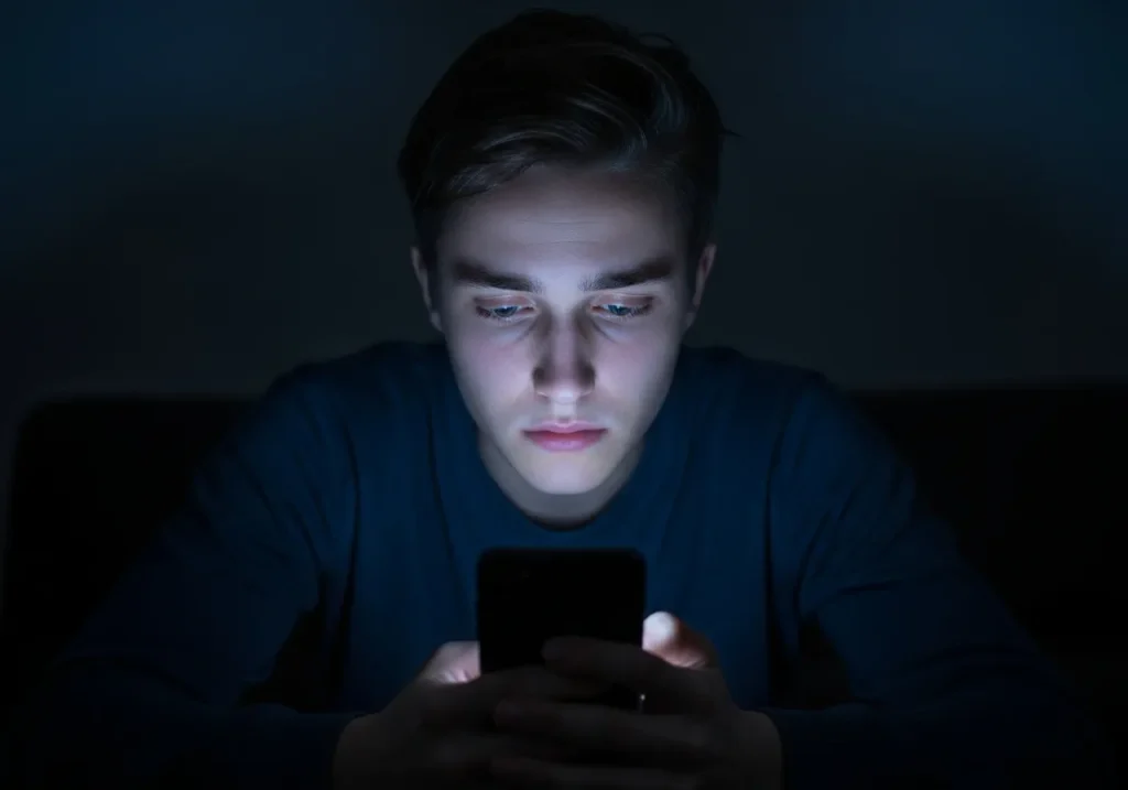Młody mężczyzna siedzi w ciemnym pokoju i wpatruje się w ekran smartfona, którego zimne światło oświetla jego twarz. Wygląda na zamyślonego i przygnębionego.